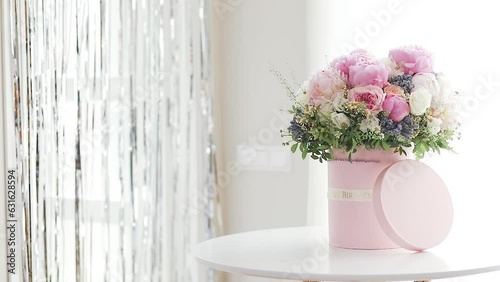 An elegant bouquet of fresh roses, peonies, and daisies in a pink box, beautifully arranged and placed by a light-filled window.
