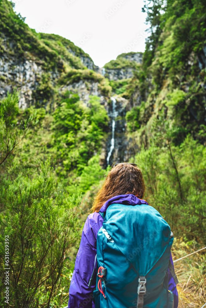 Naklejka premium Female tourist with backpack watching green overgrown Risco waterfall from the distance. 25 Fontes Waterfalls, Madeira Island, Portugal, Europe.
