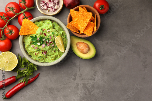 Bowl of delicious guacamole, nachos chips and ingredients on grey table, flat lay. Space for text