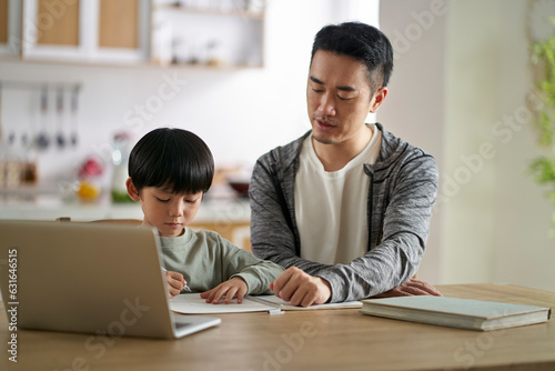 young asian father sitting at kitchen table at home helping son with homework