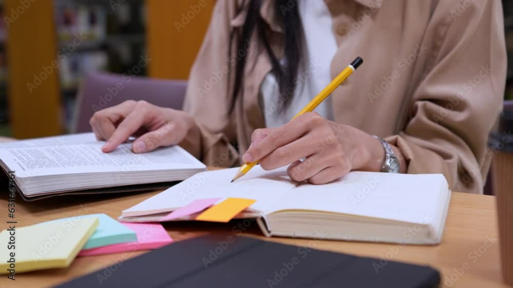 Asian women student sits in library, prepare for exam and making notes in notebook preparing for college test and finding information with a serious face in college library. Asian women student sits in library, prepare for exam and making notes in notebook preparing for college test and finding information with a serious face in college library.