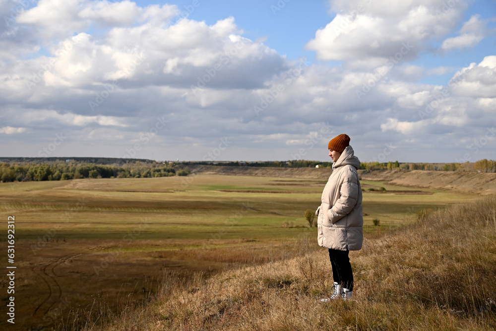 A girl in a light jacket and a brown hat looks at the autumn landscape