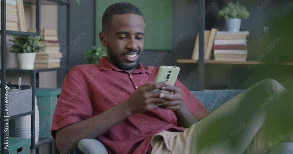 Joyful student using smart phone texting in social media sitting in armchair in apartment. Modern technology and online communication concept. Joyful student using smart phone texting in social media sitting in armchair in apartment. Modern technology and online communication concept.