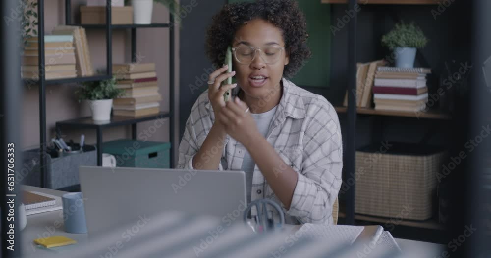 Young African American woman freelancer talking to client on mobile phone and using laptop in home office. Communication and professional occupation concept. Young African American woman freelancer talking to client on mobile phone and using laptop in home office. Communication and professional occupation concept.