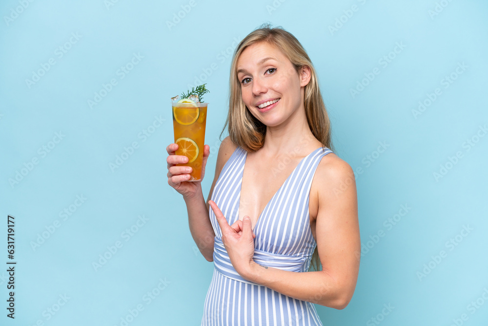 Young blonde woman wearing swimsuit holding a cocktail isolated on blue background and pointing it Young blonde woman wearing swimsuit holding a cocktail isolated on blue background and pointing it