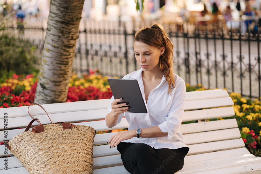 Portrait of attractive young lady in forma outfit sitting on bench in centre of city and using tablet.