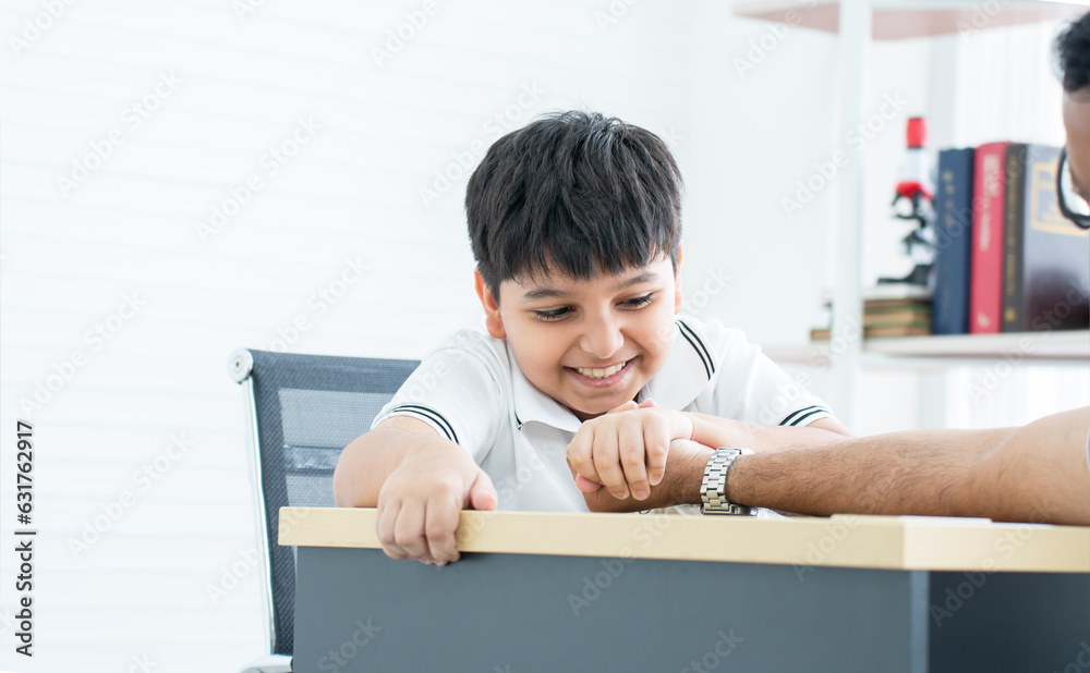 Naklejka premium Little handsome Indian child son and his father, have fun playing, competing in arm wrestling together on table at home. Cute boy win his dad. Education, Family relationship, Activity Concept
