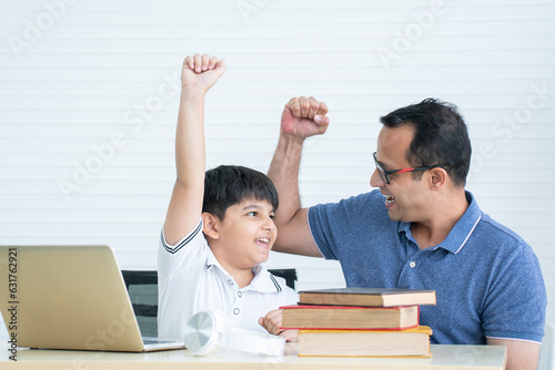 Cheerful indian family, child son with father have fun raising fist hands up together while doing homework, using laptop and books at home, single father with little boy sitting at home