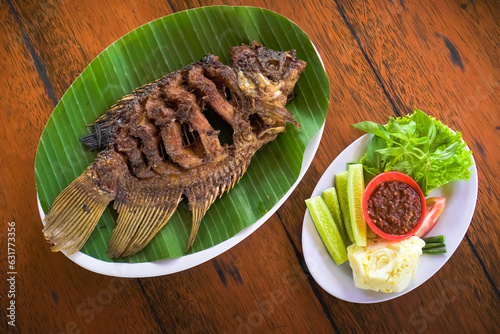 Ikan Gurame Bakar or grilled freshwater fish with chilli sauce and vegetable. Serving on white plate on wooden table. Indonesian food and cuisine. Delicious roasted Gouramy. photo