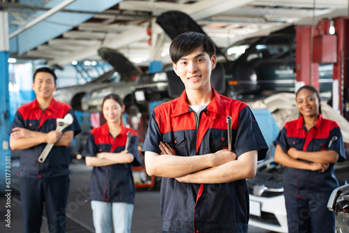 Selective focus of a young Asian male mechanic in uniform, standing to hold a wheel spanner with arms folded and smiling at camera with a blurred mechanic team standing in the background in the garage