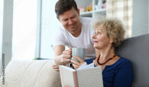 Portrait of smiling man giving cup of tea to mom. Woman sitting on sofa and reading book. Family spending time together. Warm relationship concept