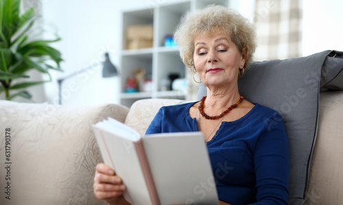 Portrait of adult woman reading interesting book. Aged curly-haired grandmother sitting on sofa at home. Smiling older grandma enjoying free time. Retirement vacation concept #631790378
