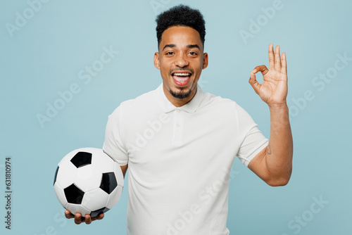 Young smiling happy man fan wear basic t-shirt cheer up support football sport team hold in hand soccer ball show ok okay gesture watch tv live stream isolated on plain pastel blue background studio.