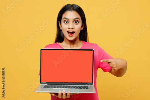 Young surprised IT Indian woman wearing pink t-shirt casual clothes hold use work point on laptop pc computer with blank screen workspace area isolated on plain yellow background. Lifestyle concept.