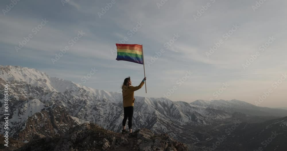 Young woman runs to the mountain peak with rainbow LGBT flag and proudly raises it up