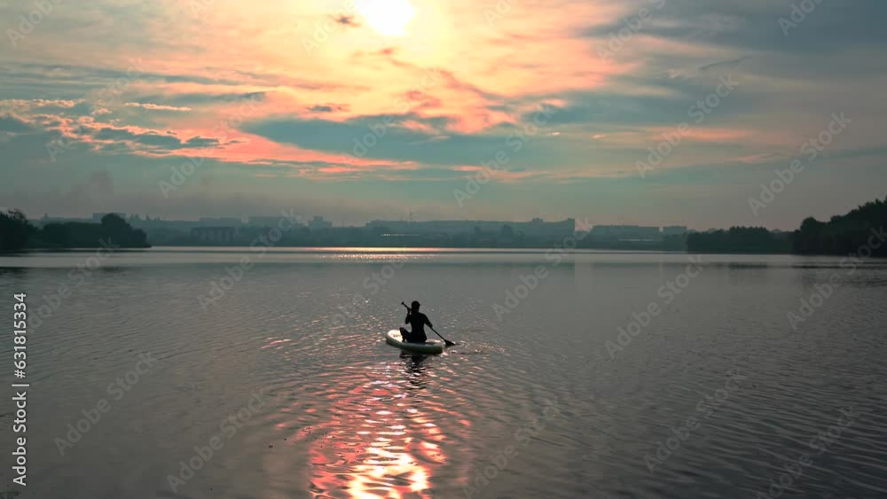 Young woman sailing on a supboard on beautiful calm lake with crystal clear water against the backdrop of the city and the cloudy sky with the setting sun. SUP stand up paddle board and girl on river