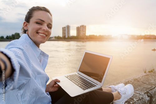 Young urban woman taking a selfie while working on laptop outdoors in front of river and cityscape on background.