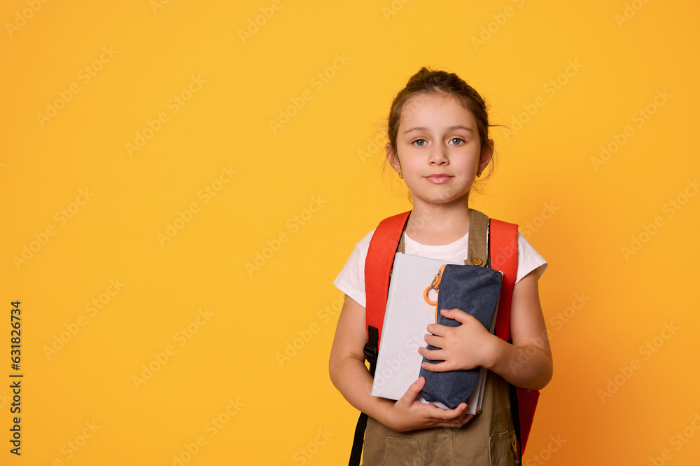 Elementary age child, primary school student girl carrying backpack, smiles cutely looking at camera, orange background Elementary age child, primary school student girl carrying backpack, smiles cutely looking at camera, orange background