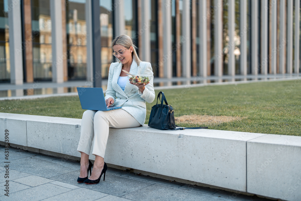 Cheerful happy young business woman looking and texting on laptop and holding salad on a break. Cheerful happy young business woman looking and texting on laptop and holding salad on a break.