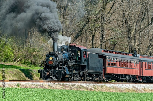 A steam passenger train moving slowly blowing lots of black smoke and white steam on a sunny day.