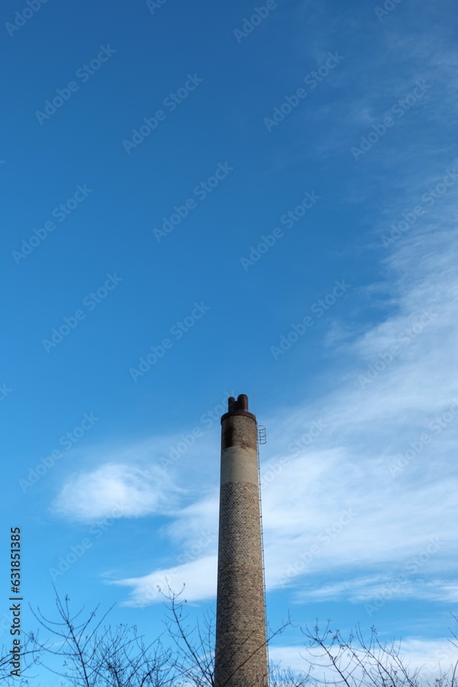 Vertical shot of a chimney with blue sky and clouds and some trees have no leaves