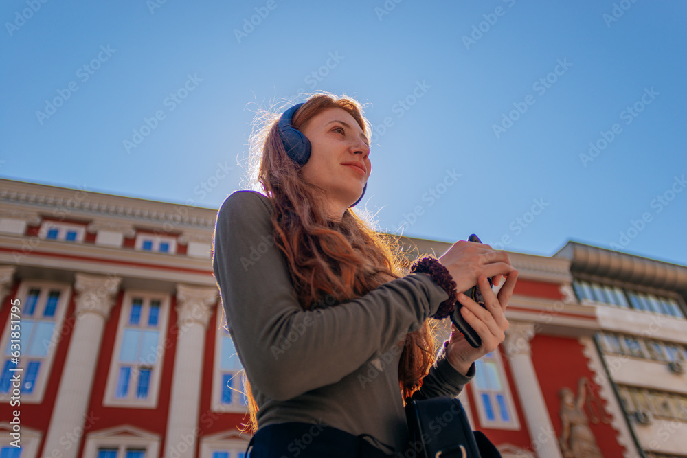 Fototapeta premium Low angle view shot of lovely ginger girl with headphones on, holding a phone and looking away