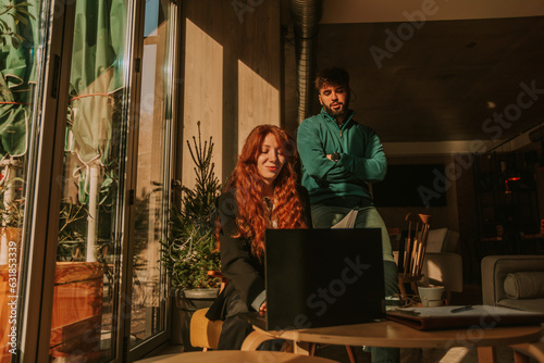 Handsome male person standing behind his female ginger colleague and watching her working on the lap top. He is giving suggestions