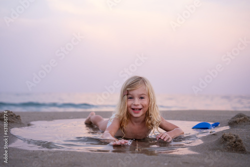 Funny smiling three years old girl lying in puddle on sandy beach , laughing, having fun at seaside looking at camera. Montessory, sensory skills development concept. Copy space. photo