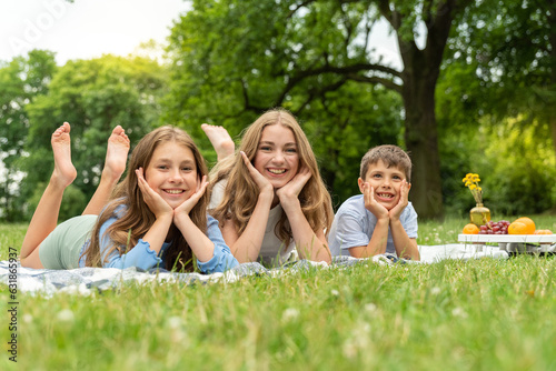 Portrait of happy three children lying in the park on a blanket on the grass. Two blond girls and one little boy prop their heads up with their hands and smile for the camera. Picnic at day off
