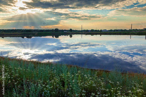 Fototapeta Naklejka Na Ścianę i Meble -  A cloudy sky with the rays of the sun breaking through the clouds and falling on a meadow, a pond and buildings visible in the distance. The wetlands near small town Rubizhne, Lugansk region, Ukraine.