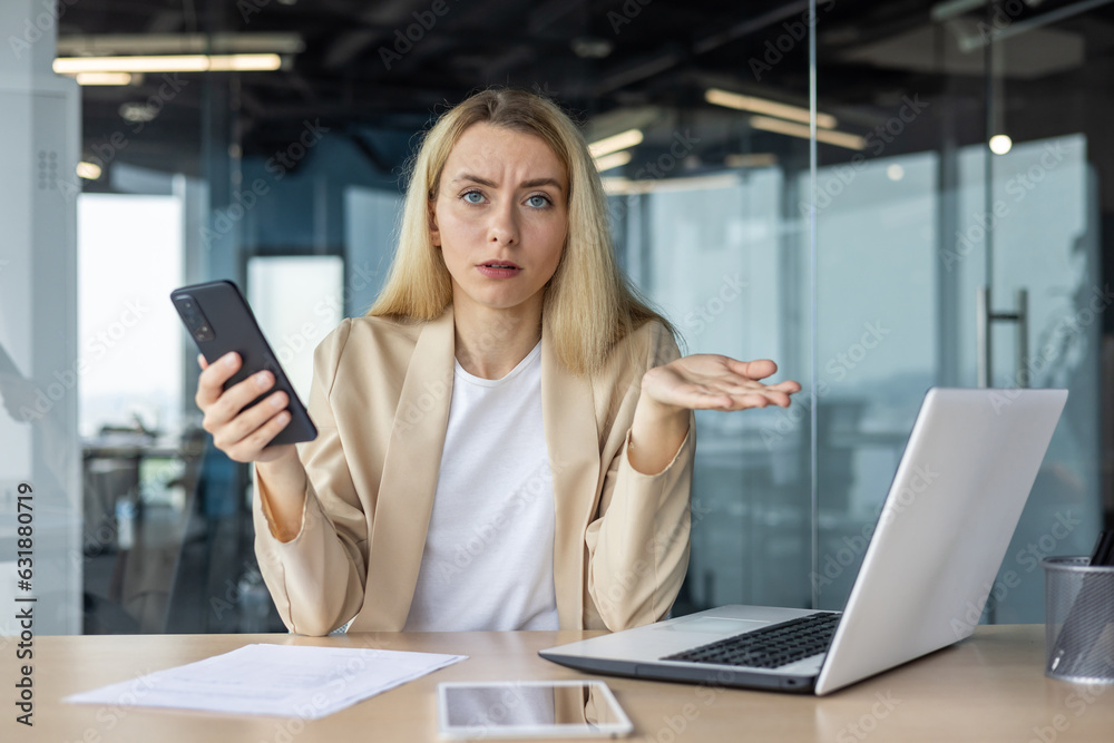 Portrait of a young business woman who is disappointed and upset sitting in the office at the table, holding the phone in her hands and spreading her hands in frustration Portrait of a young business woman who is disappointed and upset sitting in the office at the table, holding the phone in her hands and spreading her hands in frustration