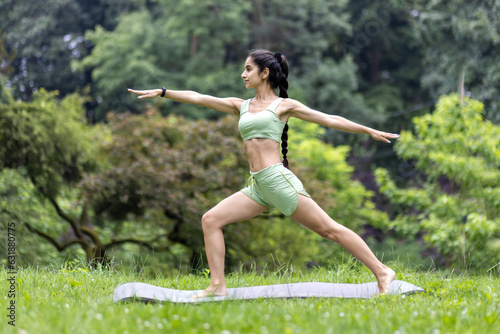 Indian young girl doing yoga outdoors, standing on a mat in a green dress, doing exercises and stretching
