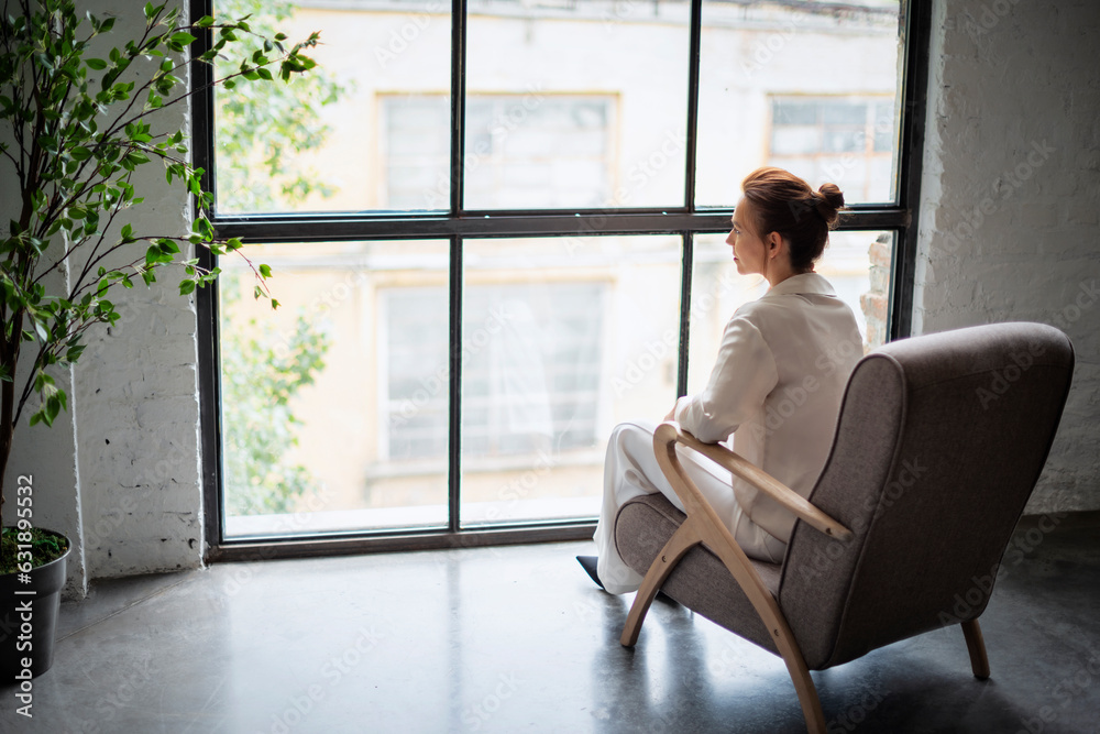 Full length of a woman sitting with her back turned in an armchair and looking thoughtfully out of the window. Full length of a woman sitting with her back turned in an armchair and looking thoughtfully out of the window.