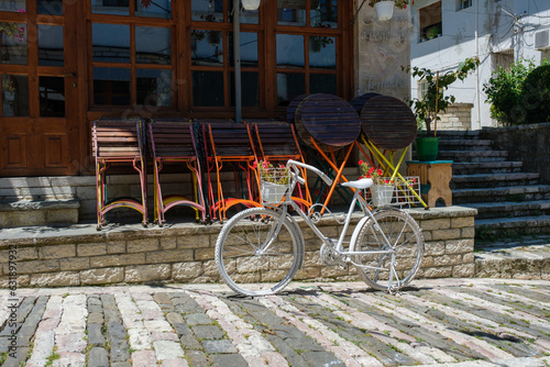 A white vintage bicycle parked on a cobblestone square in front of a rustic café or restaurant with stacked wooden chairs outside, bathed in warm Mediterranean sunlight. The charming scene captures th