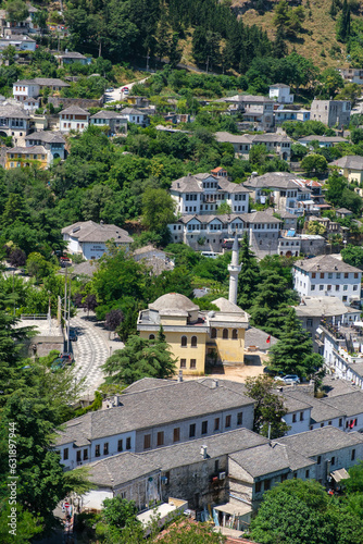 Aerial view of Gjirokaster, Albania, with a mosque, minaret, and traditional stone buildings nestled among lush green trees in summer.