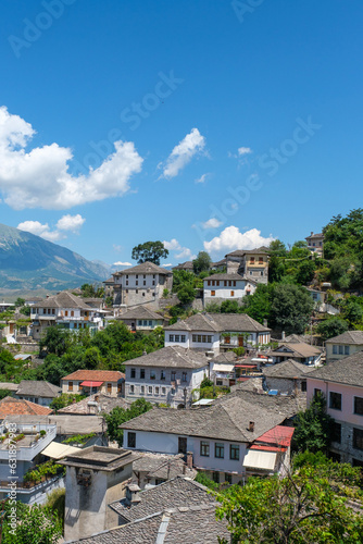 Panoramic view of Gjirokaster, Albania, with traditional stone rooftops, hillside houses, and dramatic mountains under a blue summer sky.