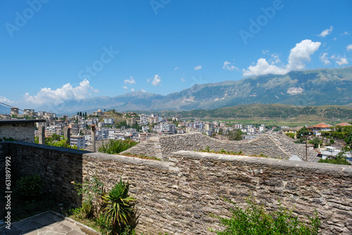 Panoramic view over Gjirokaster, Albania, with traditional stone rooftops, the city below, and dramatic mountains under a blue summer sky.
