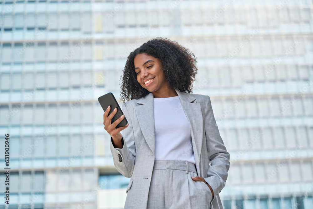 Happy young African American business woman using cell phone outdoor. Smiling lady professional holding smartphone standing at city street building looking at mobile working outside office. Happy young African American business woman using cell phone outdoor. Smiling lady professional holding smartphone standing at city street building looking at mobile working outside office.