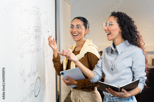 Two beautiful Hispanic female students stand near the blackboard in the classroom, point to the blackboard and rejoice at the correct solution of the formula. Girls are well prepared for college exams