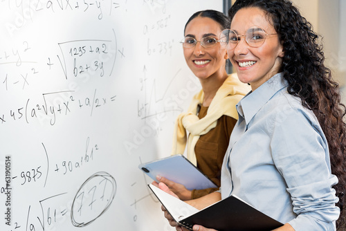 Portrait of two cute Arab high girls solving a equations on the blackboard. Girls stand near the schoolboard, prepare to enter mathematics college. Business woman in action. Smiling and looking.