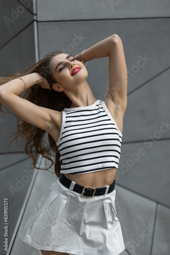 Cool fashion happy pretty woman with red lips in casual summer outfit with top and skirt stands near a geometric gray background