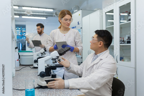 Work in the laboratory of an international team of scientists. An Asian man examines under a microscope and discusses with a female colleague. A third man in the background takes stock of the drugs.