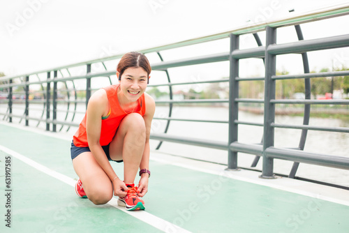 Asian woman sitting and tying running shoes before work out. Sportive female preparing running on the bridge and looking at camera.