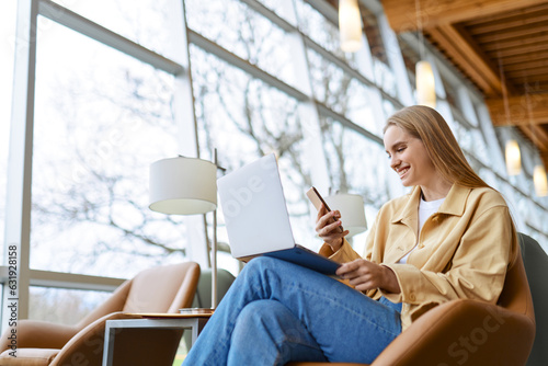 Canvas Print Happy young woman looking at cellphone, smiling girl student holding smartphone using mobile cell phone and laptop technology surfing web sitting in chair in university campus, coworking office space