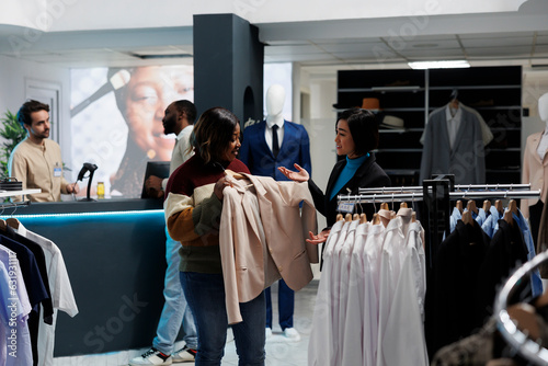 Smiling african american woman holding jacket on hanger and talking with consultant in boutique. Customer and assistant discussing fashion trends while choosing outfit in clothing store