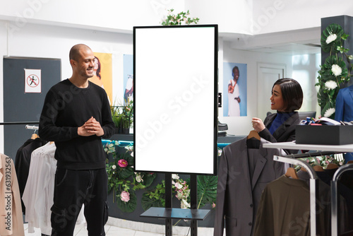 Smiling clothing store assistant chatting with arab customer while standing near blank advertising whiteboard. Shopping center asian woman employee helping client to choose apparel