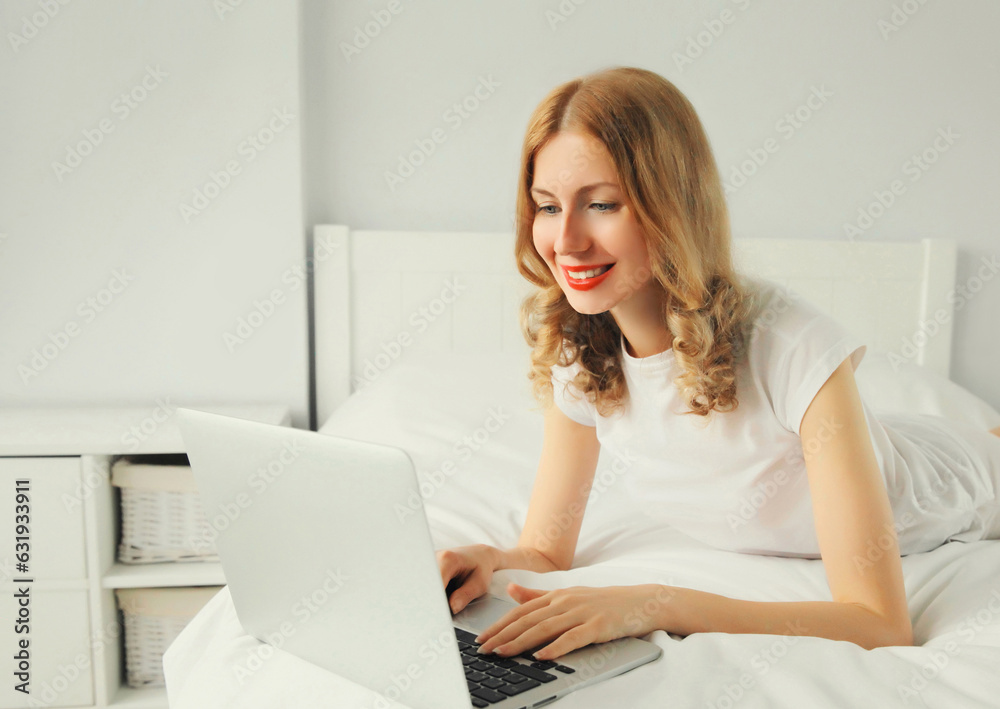 Portrait of smiling young woman working with laptop on the bed in white room at home Portrait of smiling young woman working with laptop on the bed in white room at home