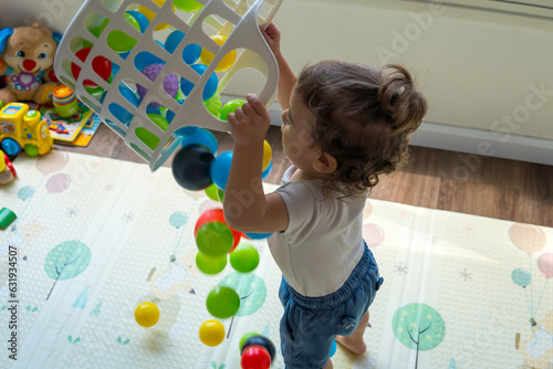 Infant baby playing with colorful plastic balls toy on a playmat. Toddler boy playing with toys at nursery, daycare, or kindergarten while standing. A child with educational toys, Early development.