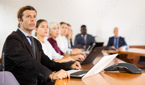 Man in suit sitting at table with his colleagues, attending meeting and using laptop.
