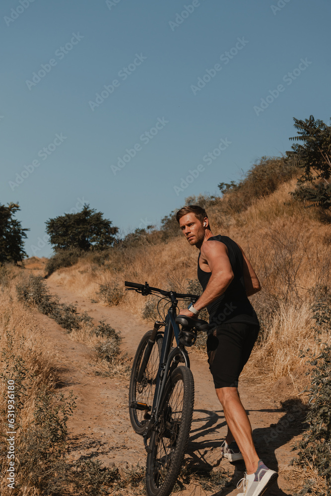 Confident young man in sports clothing pulling bicycle by mountain hill outdoors Confident young man in sports clothing pulling bicycle by mountain hill outdoors
